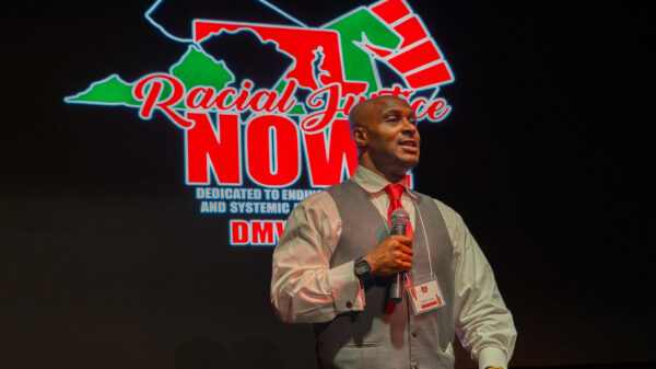 Hashim Jabar stands at a podium with the Racial Justice Now logo on a screen behind him.