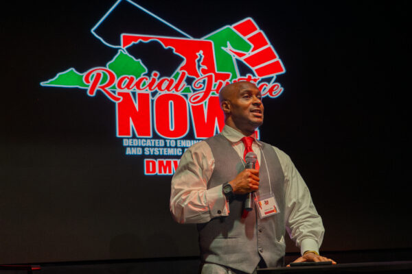 Hashim Jabar stands at a podium with the Racial Justice Now logo on a screen behind him.