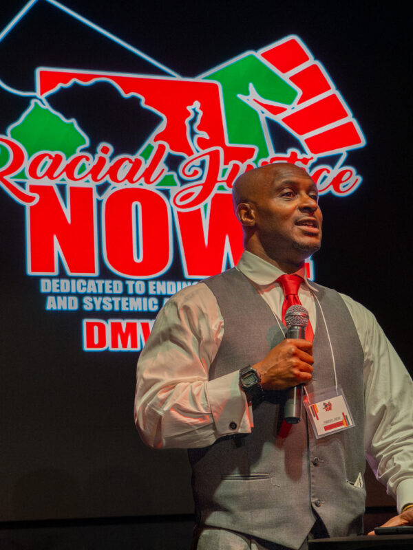 Hashim Jabar stands at a podium with the Racial Justice Now logo on a screen behind him.