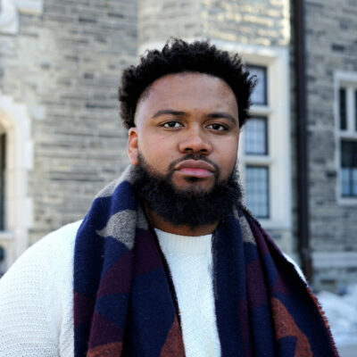 Nehemiah Bester is a Black man with short hair and a beard, and is wearing a light sport jacket, light collared shirt, and burguny tie.