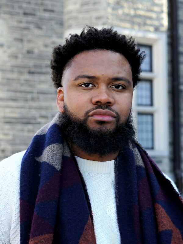 Nehemiah Bester is a Black man with short hair and a beard, and is wearing a light sport jacket, light collared shirt, and burguny tie.