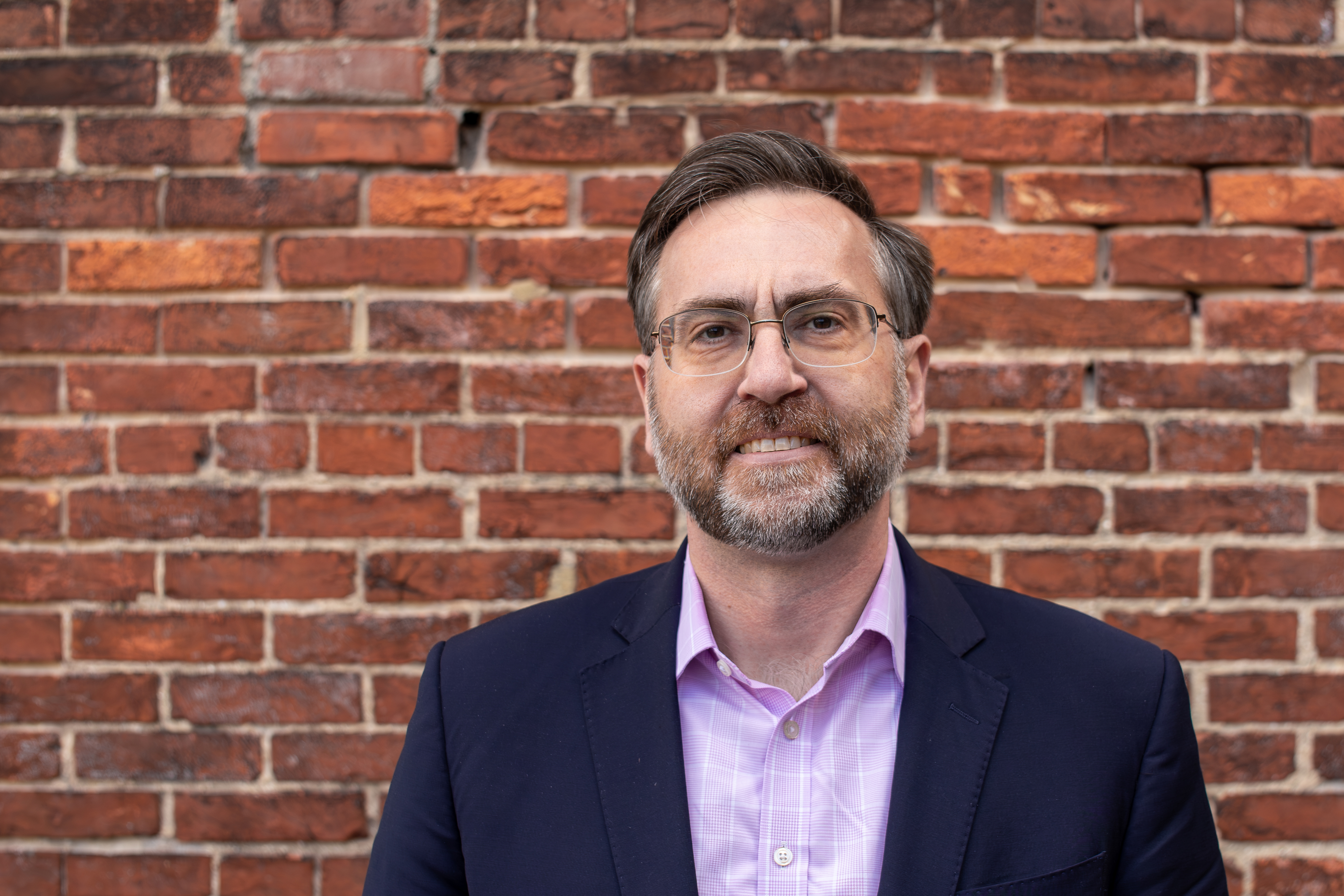 Corey Stottlemyer is standing in front of a brick wall. He is a white man with short brown and gray hair, wearing glasses and a suite.