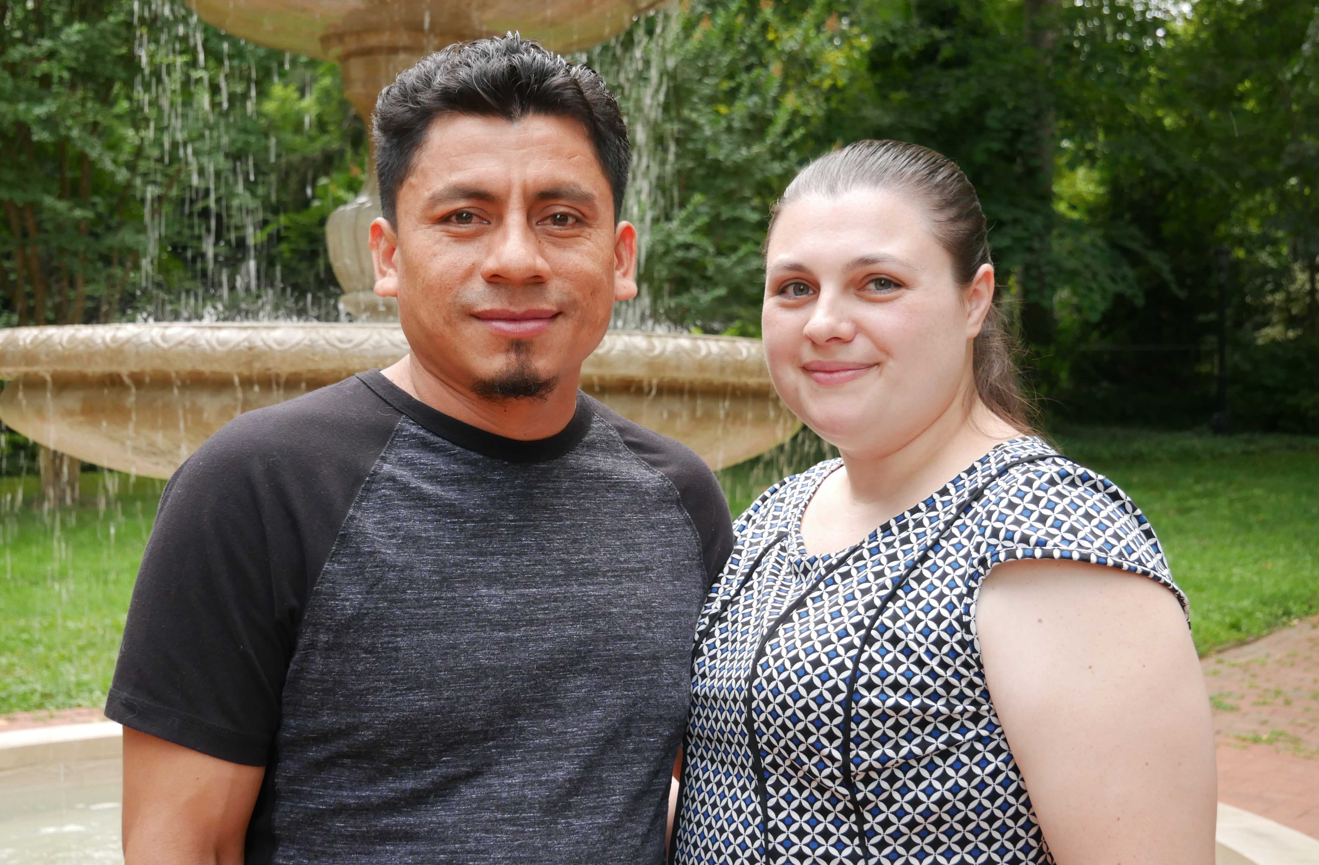 Elmer (left) and Alyse (right) Sanchez are standing in front of a water fountain with green trees behind them.
