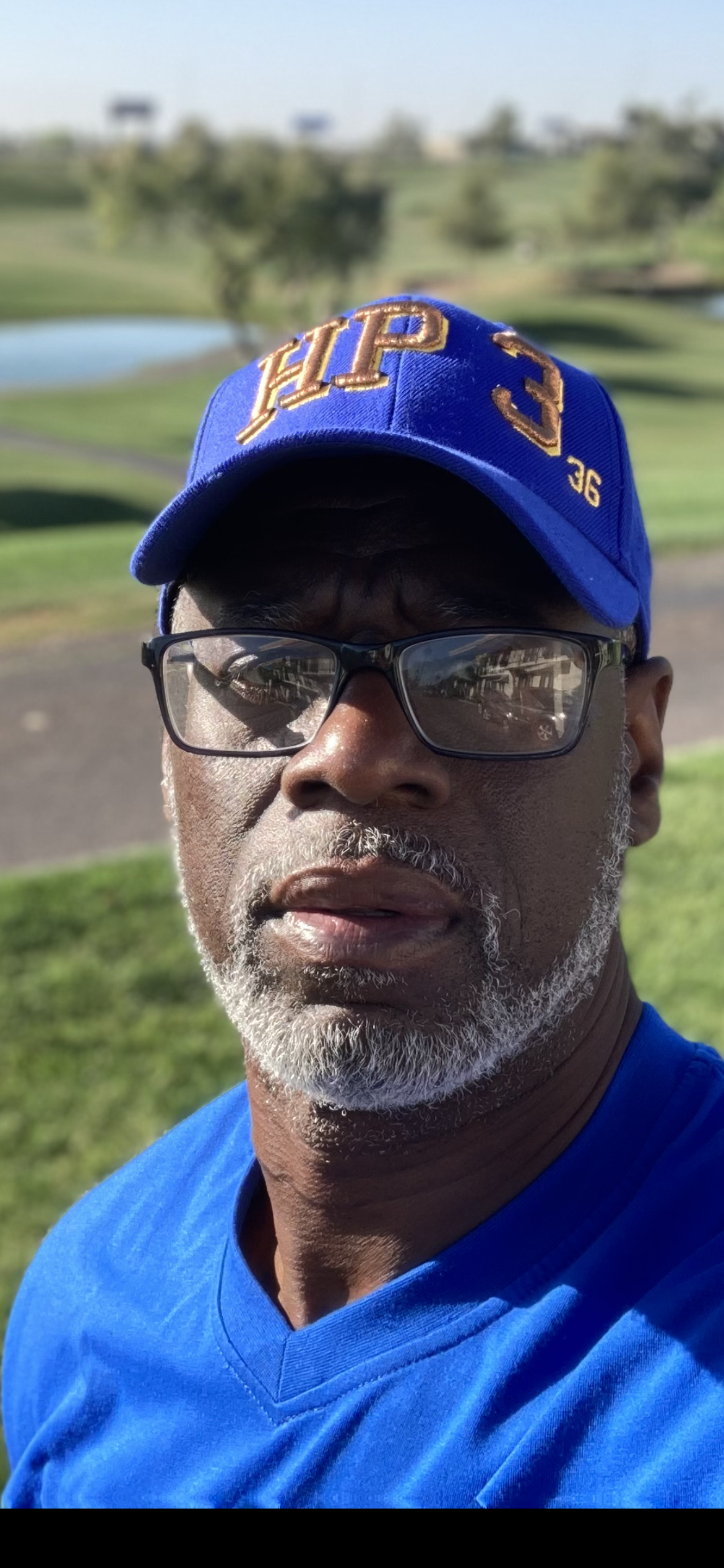 Ransom Watkins is a Black man wearing a blue baseball hat, glasses, has a short gray beard, and is wearing a blue shirt. He is standing outside.