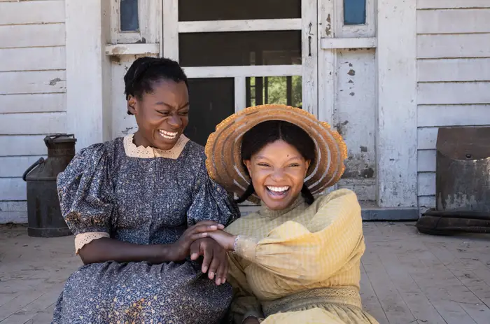 Celie and Nettiesit together on a porch in The Color Purple. A promo photo of Phylicia Pearl Mpasi as Young Celie and Halle Bailey as Young Nettie in the 2023 film.