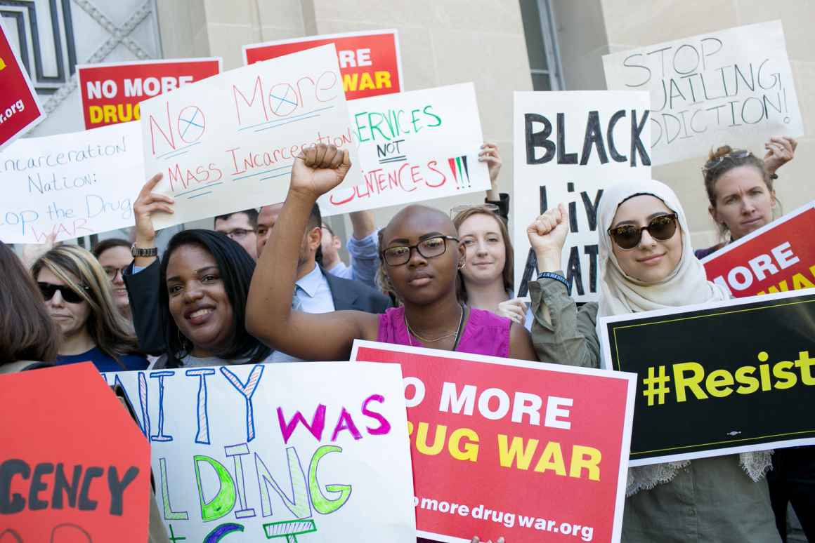 Protest with a Black person in the center with their fist raised. The signs say "no more drug war" and "Black Lives Matter" and "resist".