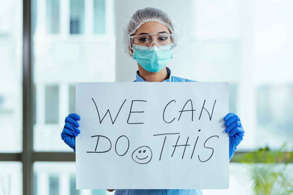 Healthcare worker that has goggles, a face mask, and gloves is holding a sign that says, "We can do this" with a smiley face.