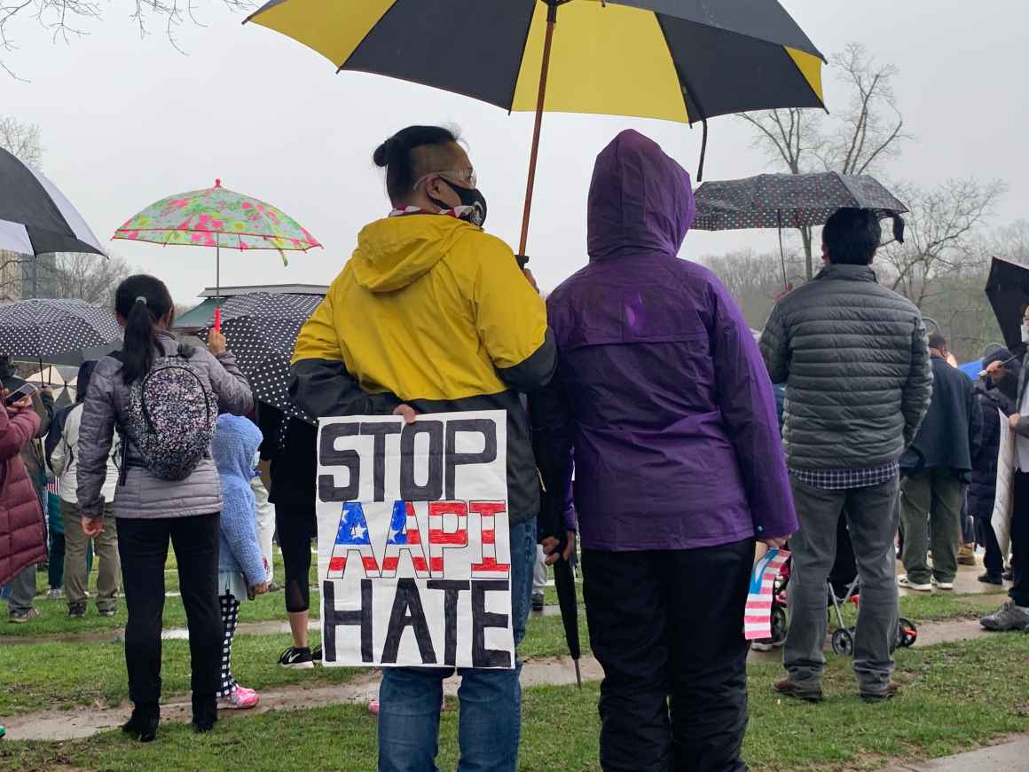 A person is standing at a rally in Columbia, Maryland, in a yellow hooded jacket, holding a yellow and black umbrella. The person is holidng a sign behind their back that says "Stop AAPI Hate". There are many other people in the picture with umbrellas.