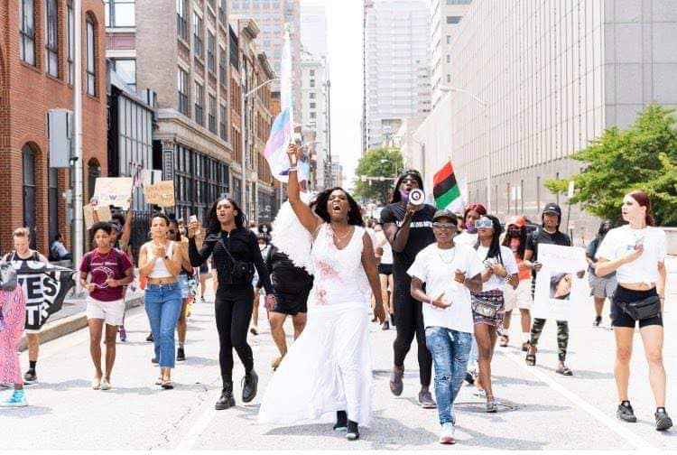 Baltimore Safe Haven photo of 2nd Black Trans Live Matter March in July 2021. Photo shows a line of people marching down a Baltimore Street. The person in the middle is holding up a trans flag.