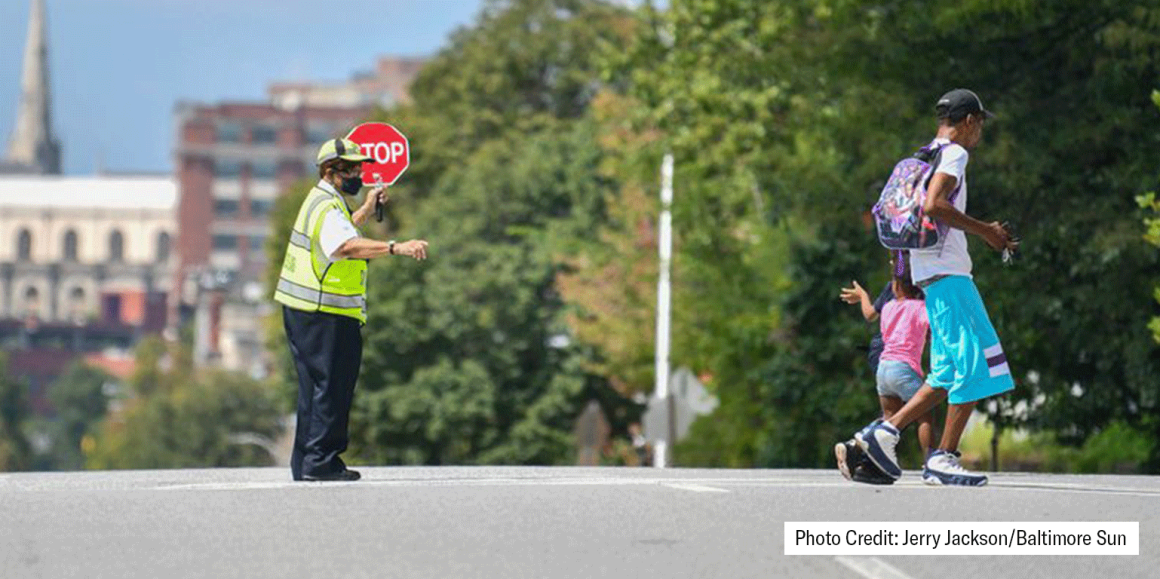 A crossing guard holds a stop sign and two students are crossing the street.