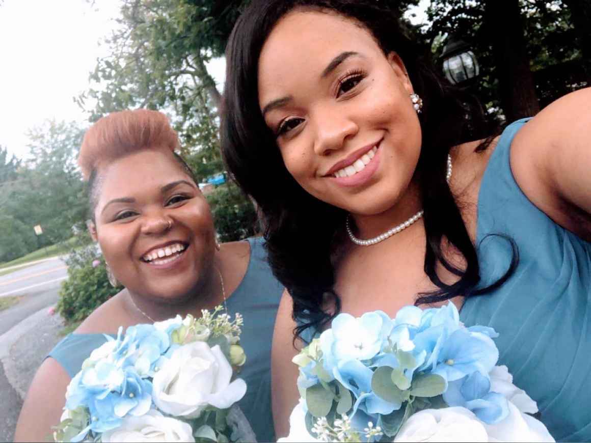 Antoinette and Alicia are sisters. They are standing next to each other wearing wedding party dresses and holding bouquets of flowers. They were bridesmaids in their older brother Harlon’s wedding in 2018.