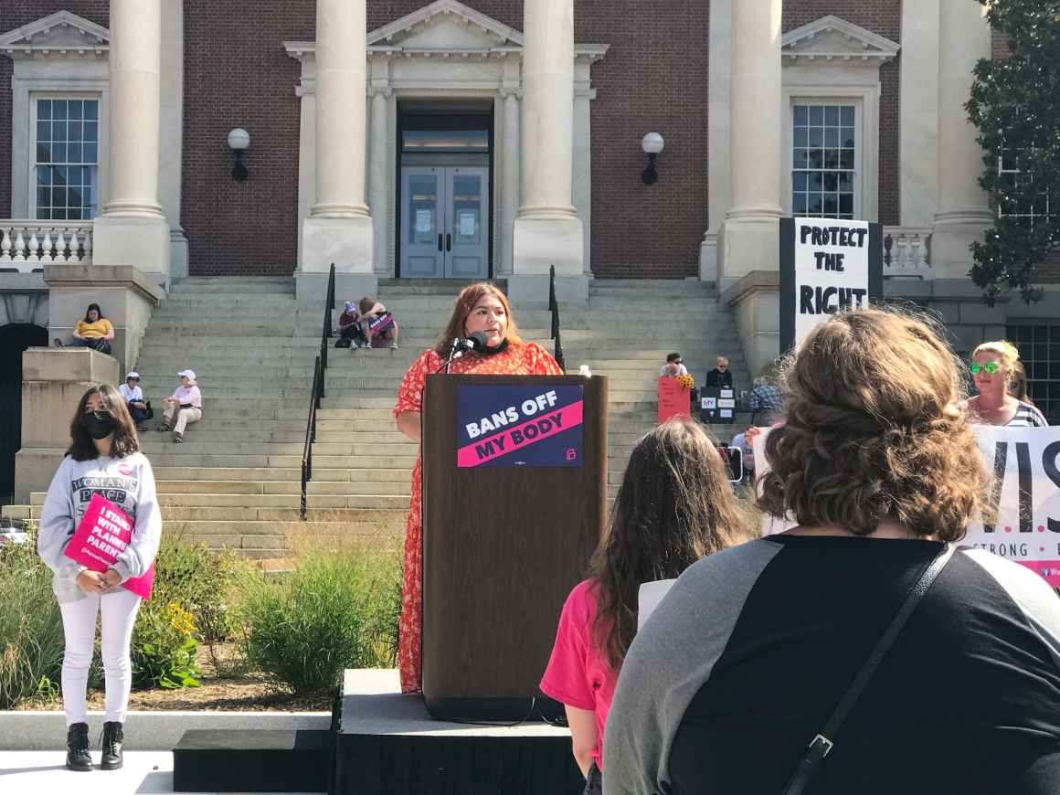 Lorena Diaz speaks at an abortion rights rally in Annapolis. She is wearing a patterned orange dress and is standing at a podium in Lawyer's Mall in front of the state house. There are people in the audience.
