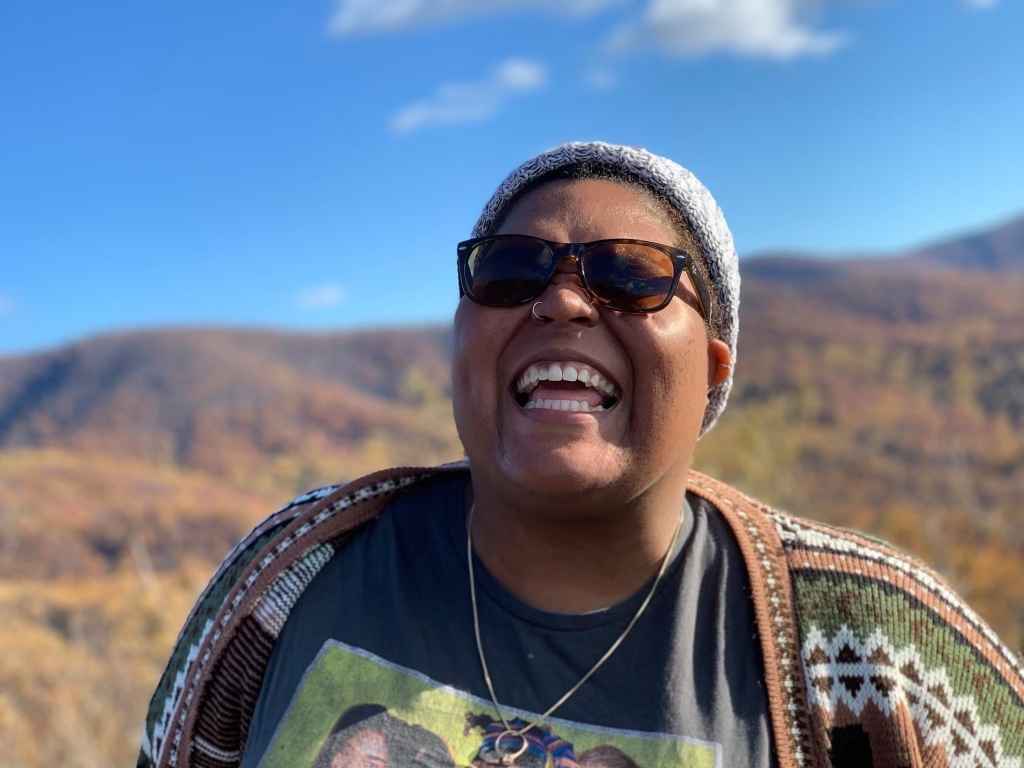 Alicia Smith is a Black, queer, non-binary person who is standing outside in front of mountains and blue sky. They are smiling and wearing sunglasses.