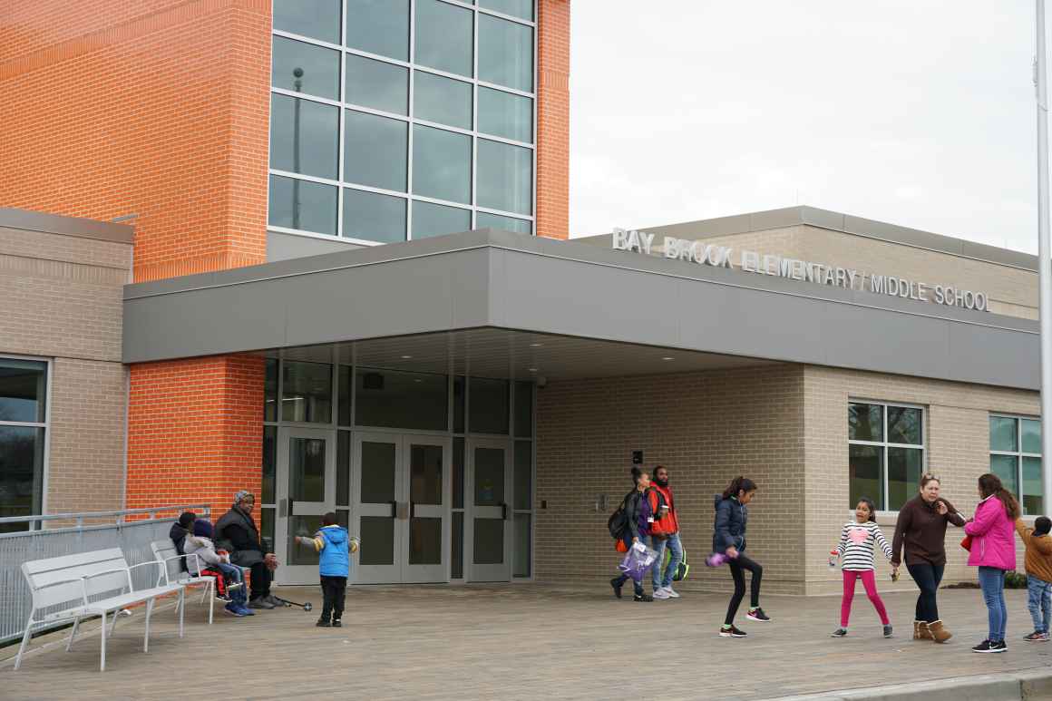 Children, parents, and school staff are standing out front of the brand new Bay-Brook Elementary / Middle School in Baltimore, MD. The are lots of windows and a colorful orange facade.