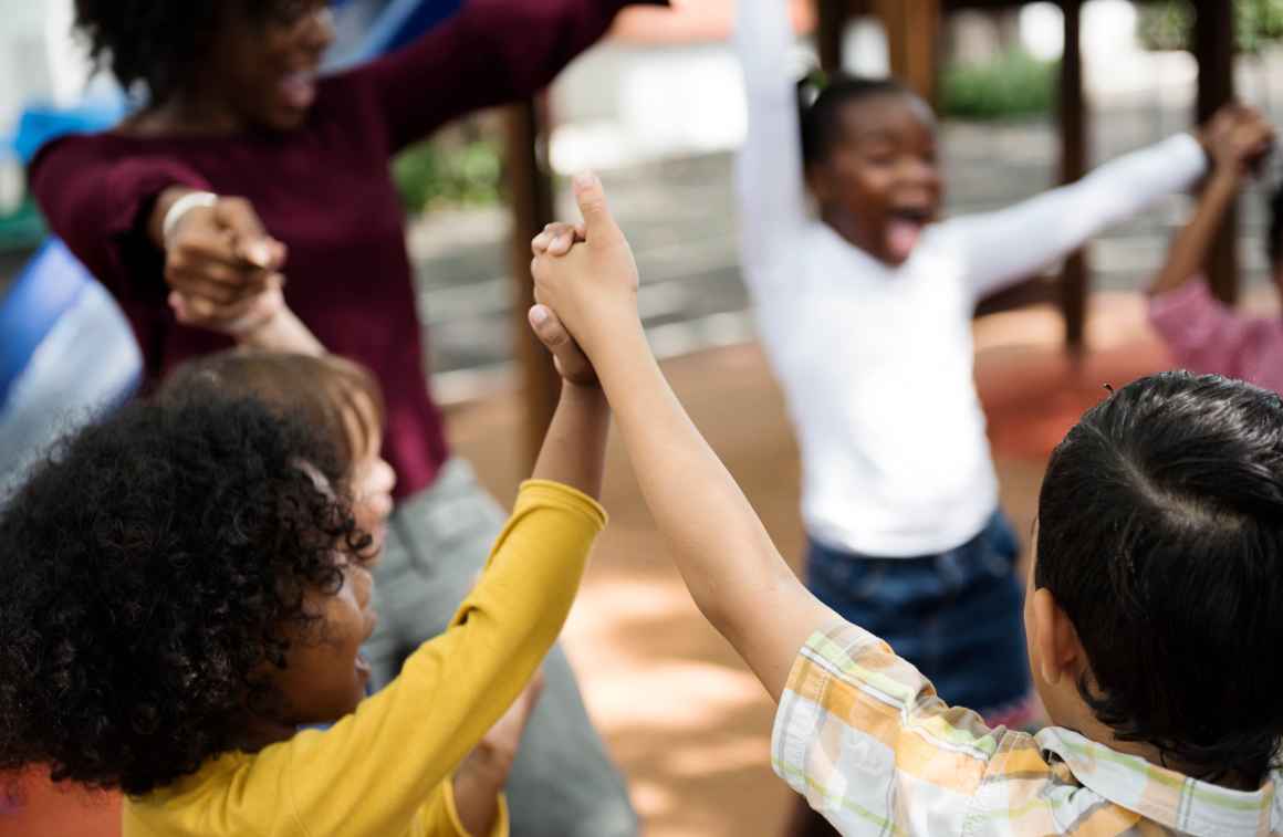 Several children are in a circle holding lifted up hands and are with an adult. They are all smiling and laughing.