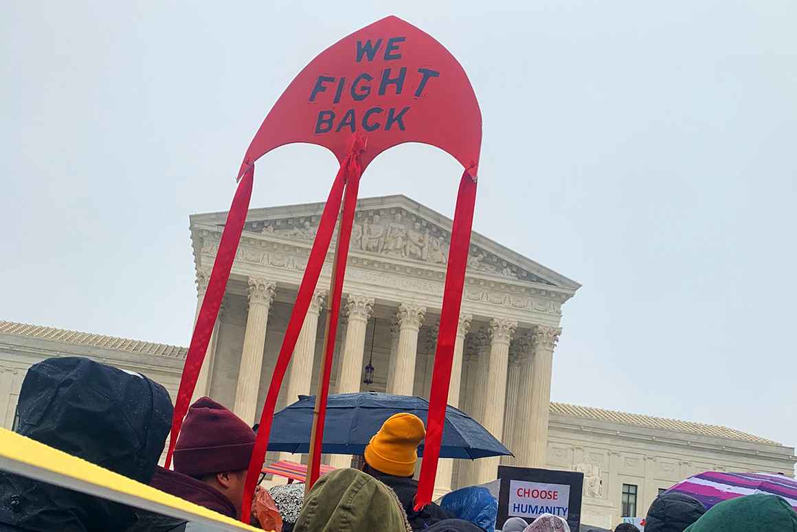 A large crowd of people are holding protest signs and gathered in the rain in front of the Supreme Court in Washington, DC. One big ride kite sign says "We fight back!"