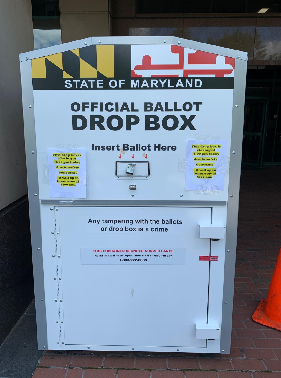 Picture of a Maryland official ballot drop box. It is a white, rectangular shaped box with the State of Maryland name and flag on it and it says Official Ballot Drop Box and insert Ballot Here. There is a small opening in the middle with three red arrows.