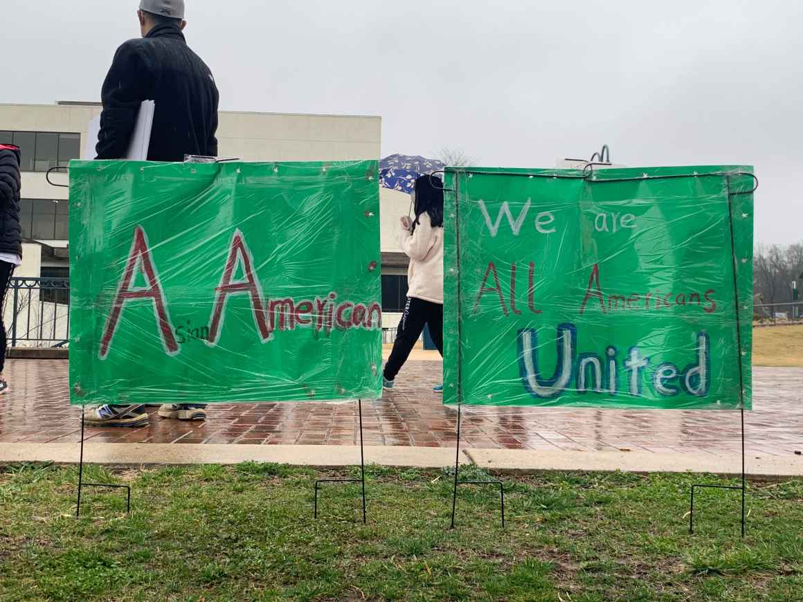 A person at the Stop Asian Hate rally in Columbia, Maryland, is standing in front of two green signs that say, "A Americans" and "We are all Americans United."