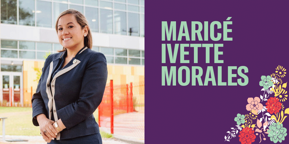 Maricé Iveltte Morales is pictured standing up and hands clasped in the front. She is a woman with medium brown skin and is smiling. She is wearing a blazer. She is standing outside in front of a building with a lot of glass windows. There are flowers.