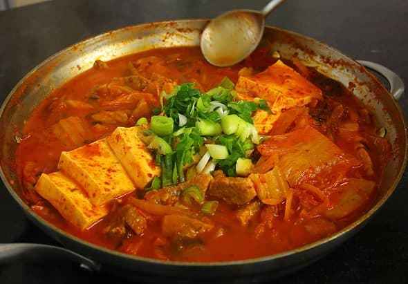 Kimchi jjigae in a metal bowl with towfu and green onions on top. The stew is a red brothy cabbage kimichi base.