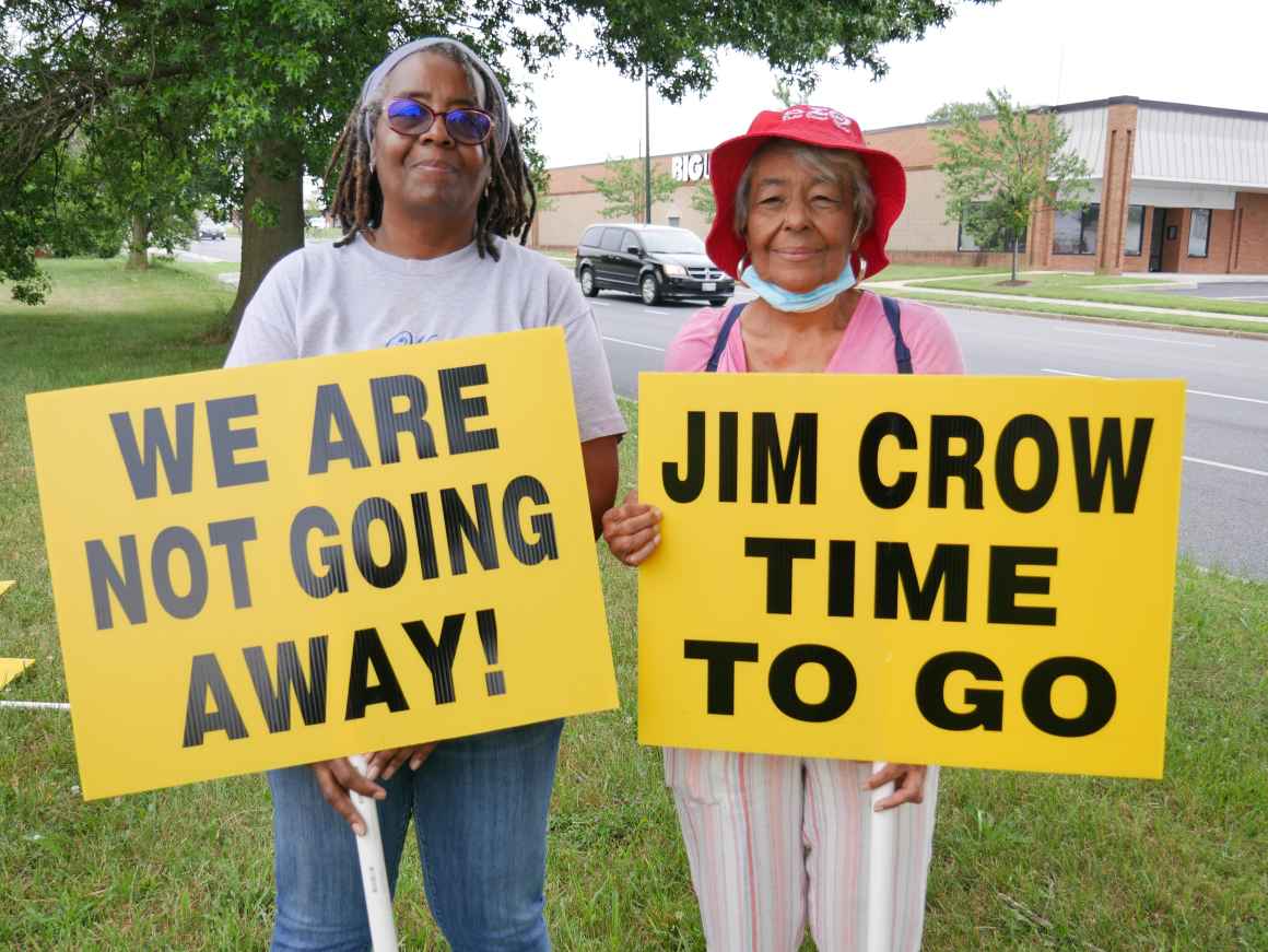 Two Black elders hold signs that say, "We are not going away!" and "Jim Crow time to go."