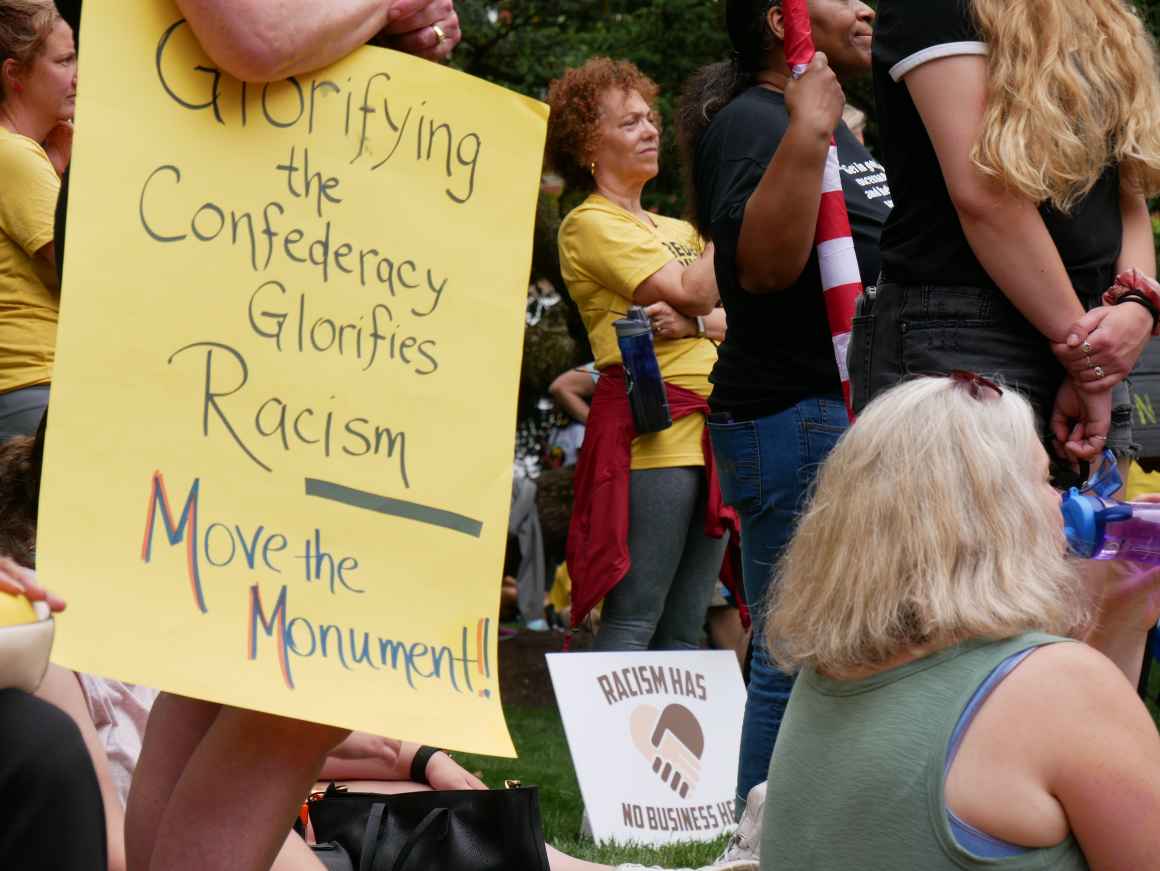 Person holds a sign that says, "Glorifying the Confederacy Glorifies Racism. Move the Monument." Another sign is visible that says, "Racism has no business here."