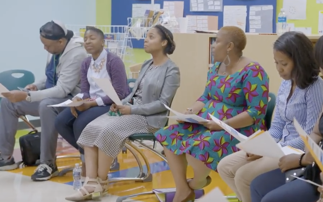 Shantay McKinily is pictured standing with six other adults, sitting in chairs at a training, learning about restorative approaches. 