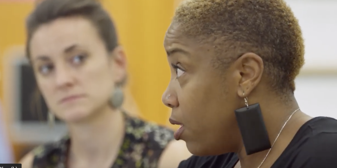 Shantay McKinily is pictured standing with a white adult. They have serious looks on their faces. The background has a yellow wall.