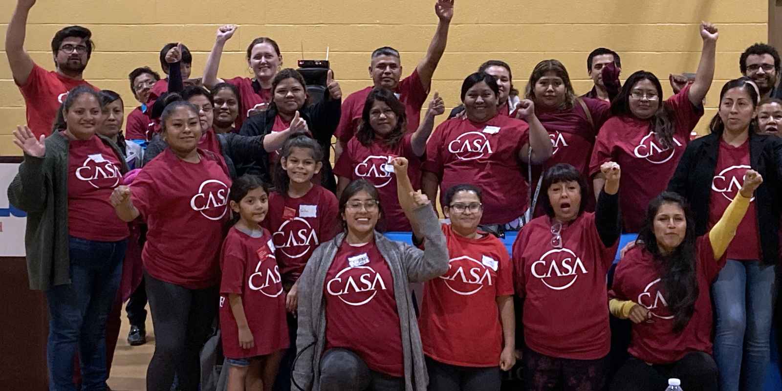 A group photo of CASA members wearing red CASA t-shirts. Several people have their first raised.