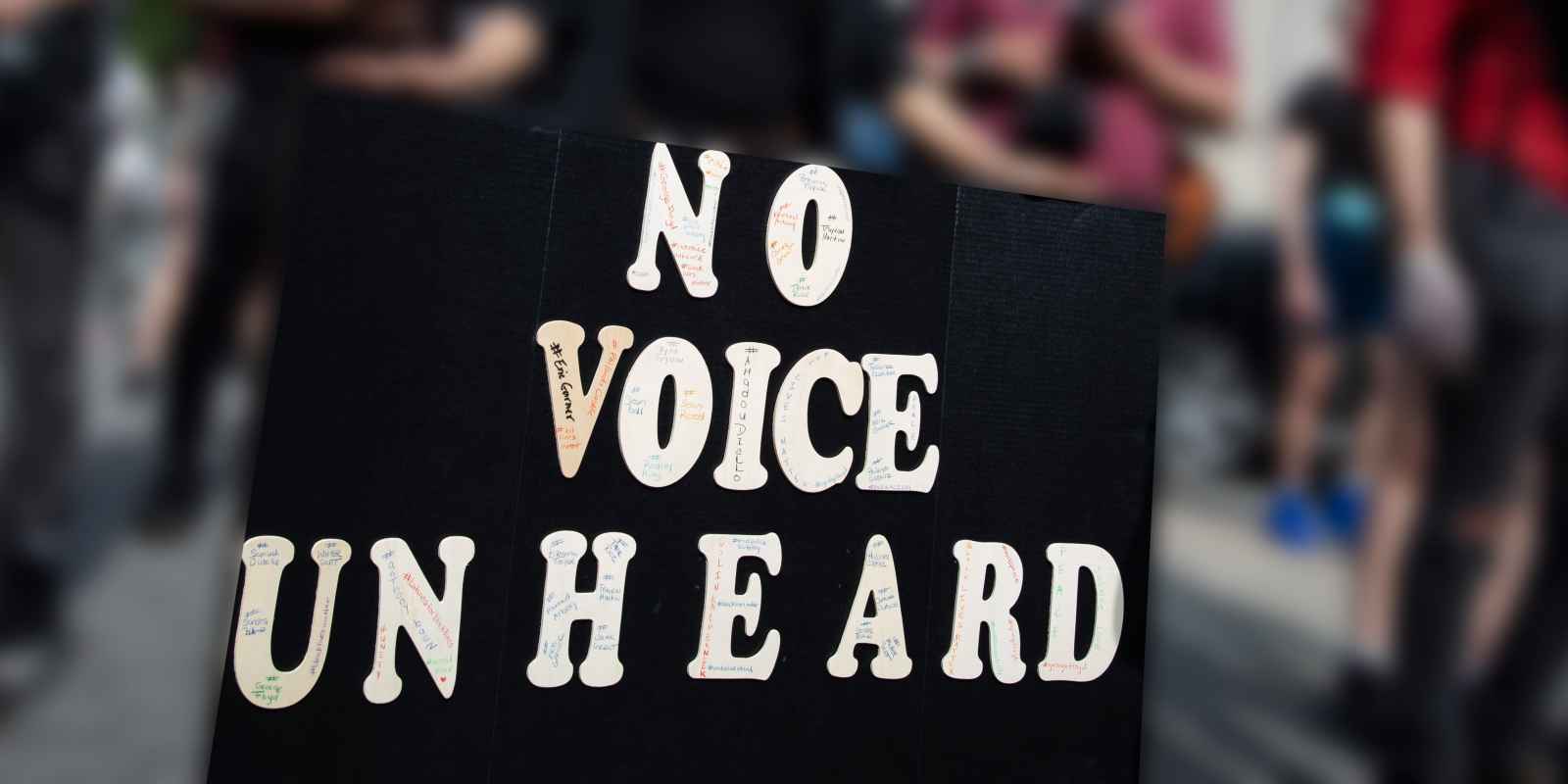A person's hands are holding a large protest sign that says, "No Voice Unheard." The background is blurred out but you can see that there are people at a protest. Courtesy credit: Molly Kaplan, ACLU. 
