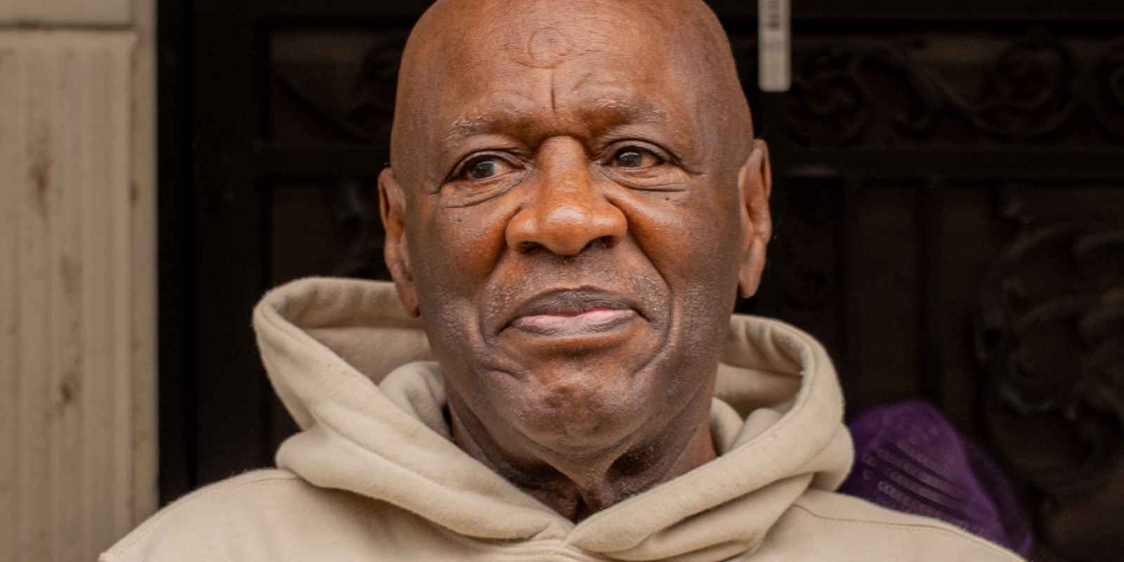 Bobby Stewart is a Black man with a beige hoodie, looking to the left, with a slight smile on his face. He is standing outside on a porch.