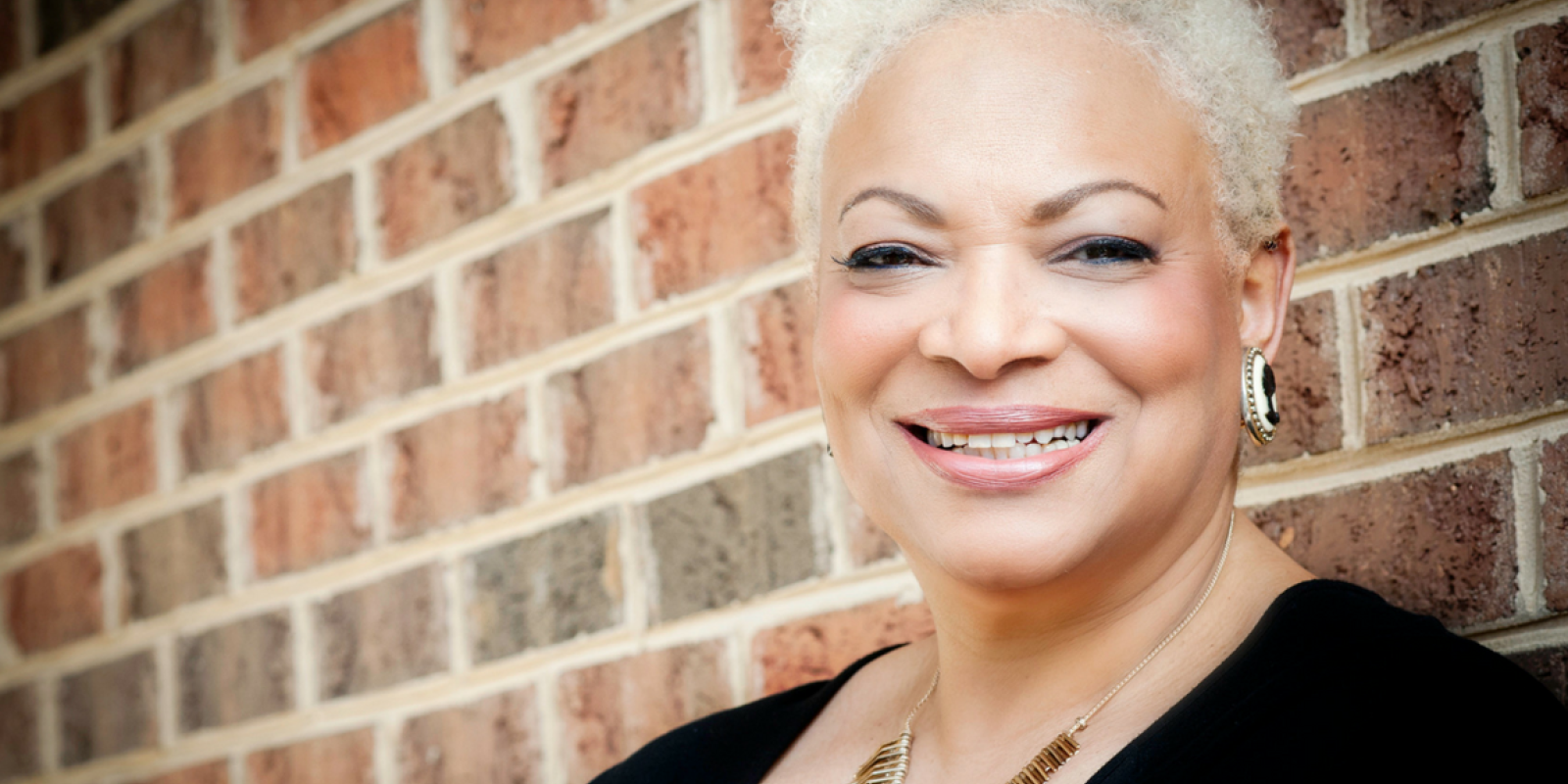 Dana Vickers Shelley is a Black woman with warm light brown skin, blond short hair, and is smiling at the camera. She is standing in front of a brick wall.