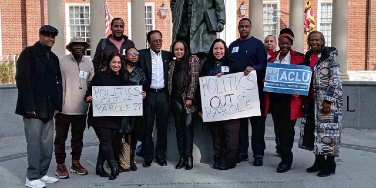 Group of advocates celebrate the veto override of parole reform in Annapolis, Maryland. They stand in front of the Thurgood Marshall statue. Some hold signs about getting politics out of parole and one person holds a sign with the ACLU-MD logo.