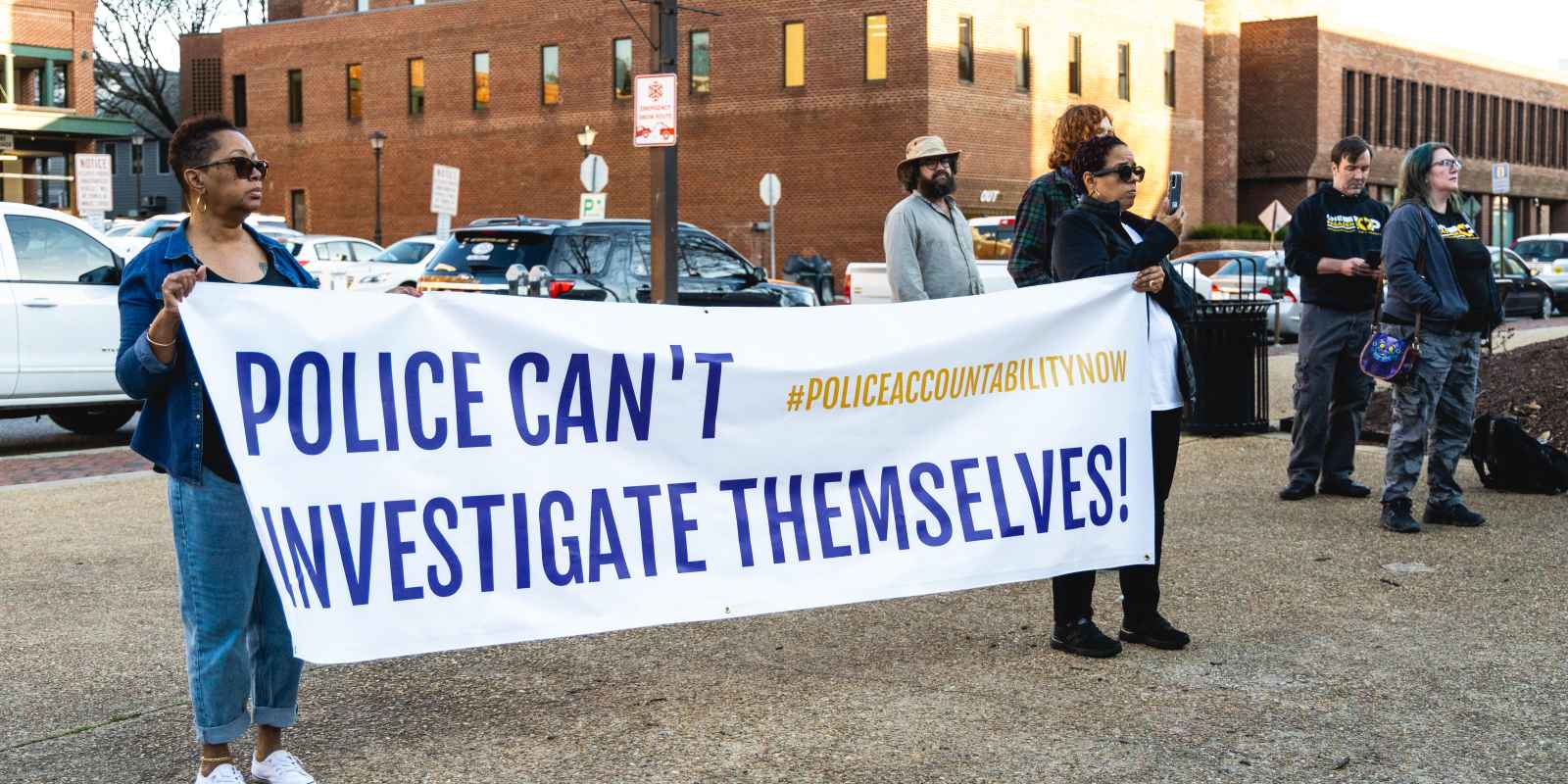 Two Black advocates hold up a banner that says, "Police Can't Investigate Themselves." They're standing outside at a rally.