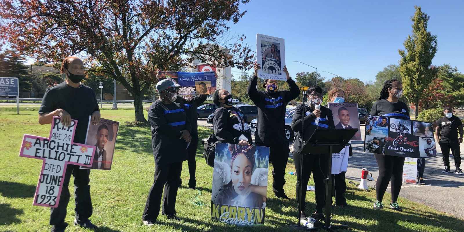 Prince George's County rally photo for mothers of Black peoole killed by police. A group of people are standing in a park holding signs with people's names who were killed.