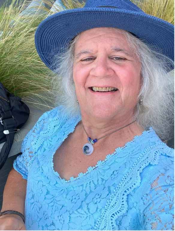 Bonnie K. Smith is a fair skinned white woman with a blue brimmed hat on, white hair, a blue lacy top and is smiling at the camera.