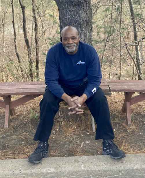 Picture of Calvin McNeill, a Black man with bald head, sitting on a bench in front of a tree.