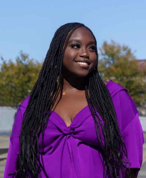 Chinyere Okonkwo is a Black woman with long dark hair. She is wearing a purple top with a v-shaped neckline. She is smiling and looking to the right. She is outside and there are trees behind her.
