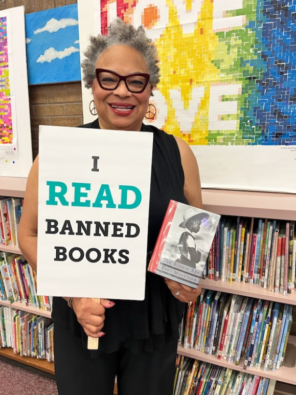 Dana Vickers Shelley is inside of a school library and holding a sign that says, "I read banned books." She is holding a copy of The Bluest Eye by Toni Morrison. Dana has short gray and black hair, is wearing cat-eye glasses and earings, and is smiling.