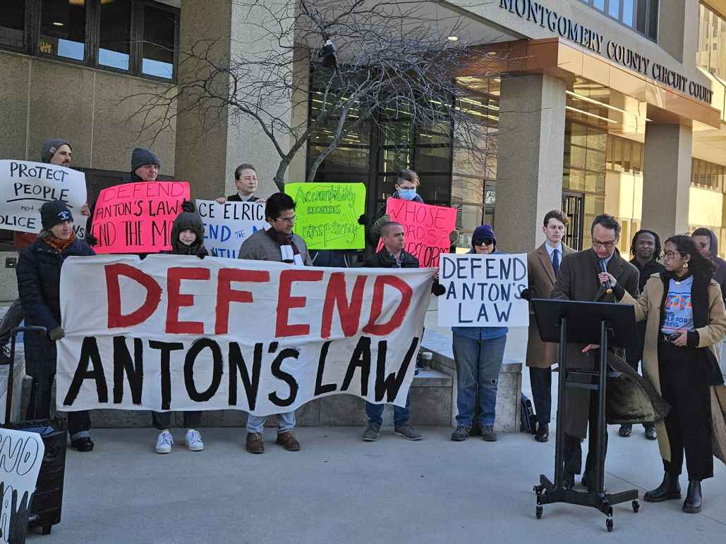 Group of advocates gather in front of the Montgomery County Circuit Courthouse at a press conference to defend Anton's Law. There is a person at a podium and about 20 advocates holding banners and signs.