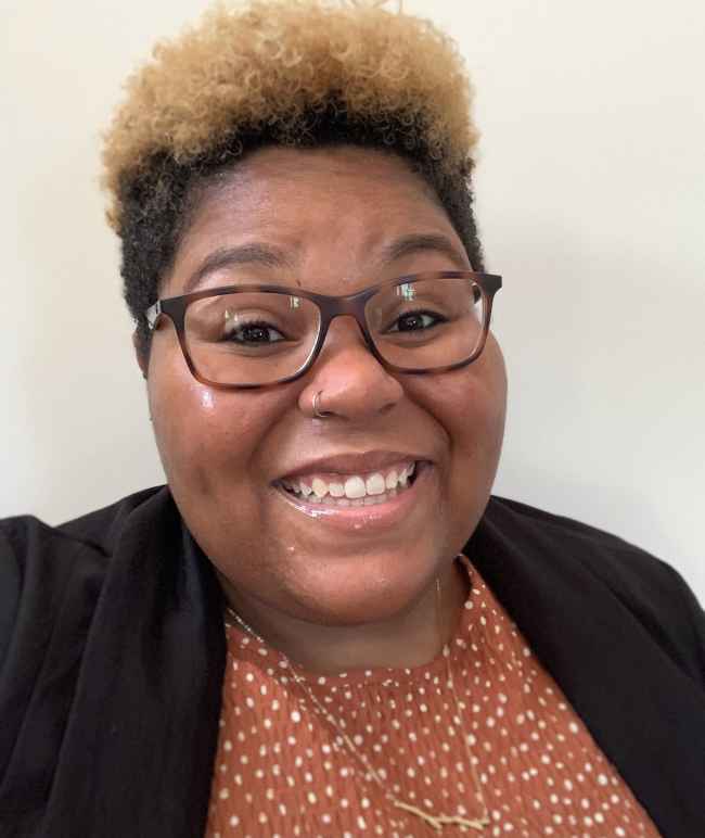 Alica Smith is a Black person, pictured with short black and bleached hair, wearing glasses, smiling, and a dark blazer over a brownish-orange shirt.