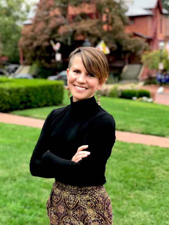 Amy Cruice is a white woman who has short medium brown hair, is wearing a black long-sleeved shirt and is standing outside in front of a yard with green grass and has her arms crossed.