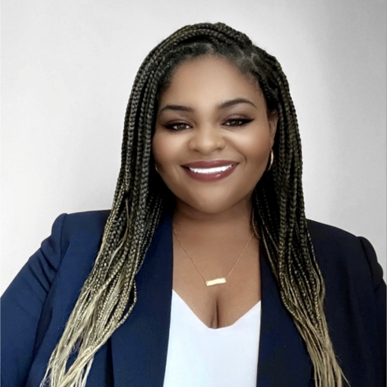 Brittanyt Christian has long braided hair, dark with light ends. She is wearing a dark blue suit jacket and white shirt. She has a warm Brown skin tone and is smiling and looking at the camera.