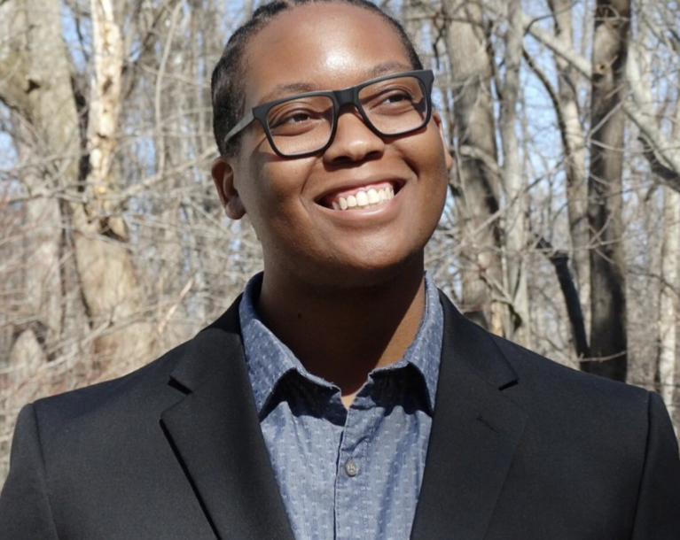 Carlos Childs is looping to the side and up and is smiling. He is a Black man and is earing dark0framed glasses. He is outside in front of trees, is wearing a black suit jacket over a blue button-up and collared shirt.