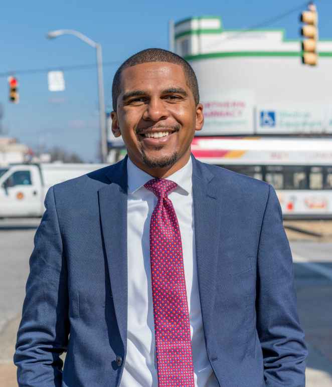 Image shows a picture of Caylin Young in a blue suit and fuschia time. He is standing on a Baltimore City street on a sunny day. He is a Black may with short hair and he is smiling in the photo.