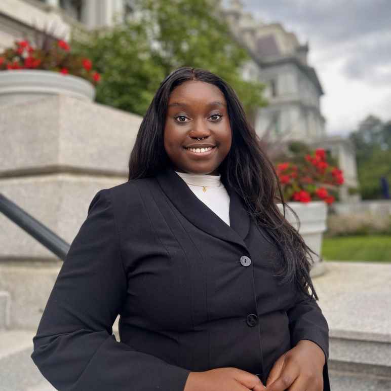 Chinyere Okonkwo is a Black woman with long dark hair. She is standing outside in front of a building with flowers and green plants. She is wearing a black blazer and is smiling at the camera.