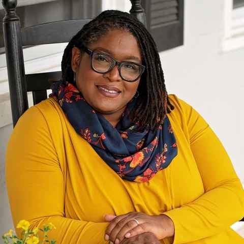 Dr. Sheila Graham is a Black woman sitting in a chair. She is wearing a yellow/orange long-sleeved shirt, a floral scarf, and glasses. Her hair is styled in lochs just above her shoulders. Her hands are clasped and she is smiling.