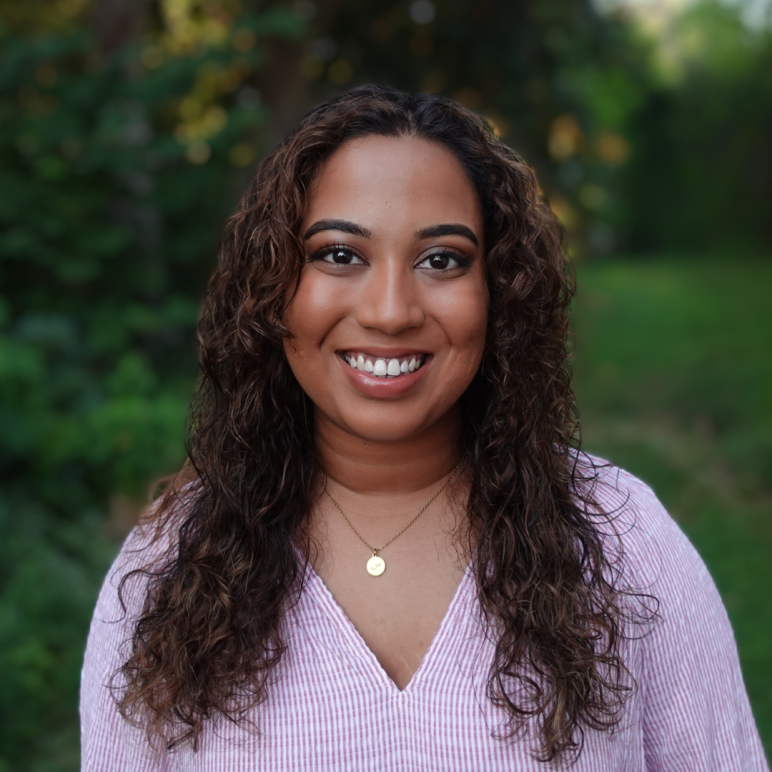 Fathima Rifkey is standing outside with trees and grassy field in the background. She has warm medium-brown skin, long curly hair, is smiling at the camera, and is wearing a pink v-neck shirt.