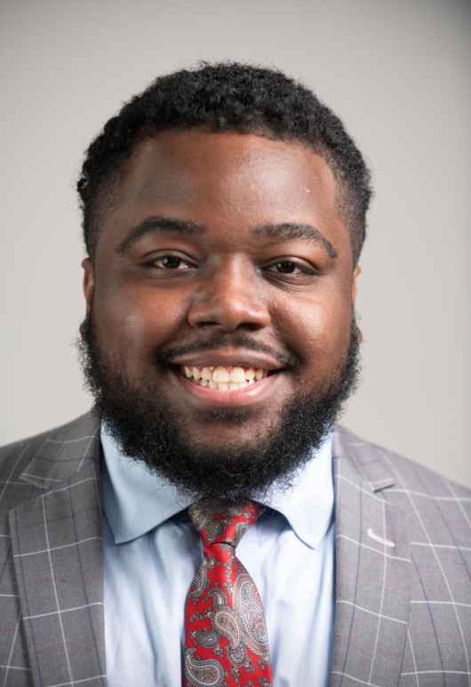 Gregory Brown is smiling at the camera. He is a Black man, has short dark hair, facial hair, and is wearing a gray plaid suit jacket, light blue button up shirt, and red patterned tie.