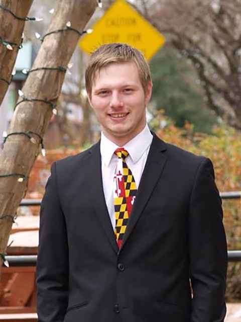 Jared is a white man with lighg colored short hair, standing in front of a tree with lights on the branches, and he is wearing a dark suit, white shirt, and Maryland flag tie. He is smiling and looking at the camera.